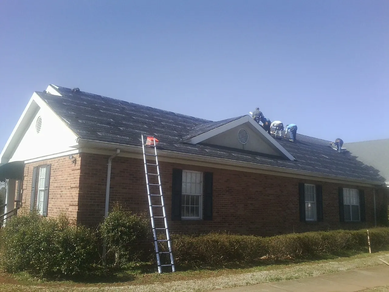 Crew tearing shingles off a brick ranch roof with a ladder set — Metal Roof Repair work by our crew in Macks Inn