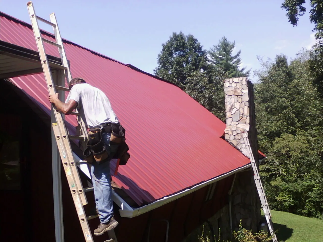 Roofer on a ladder beside a red metal roof and stone chimney — Soffit & Fascia Repair by Roofing Brothers in Macks Inn